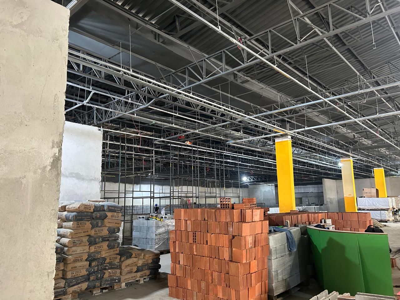 Obra da Bio Ritmo Recife em execução, com estrutura metálica da cobertura, instalações suspensas, pilares amarelos e materiais estocados no piso.