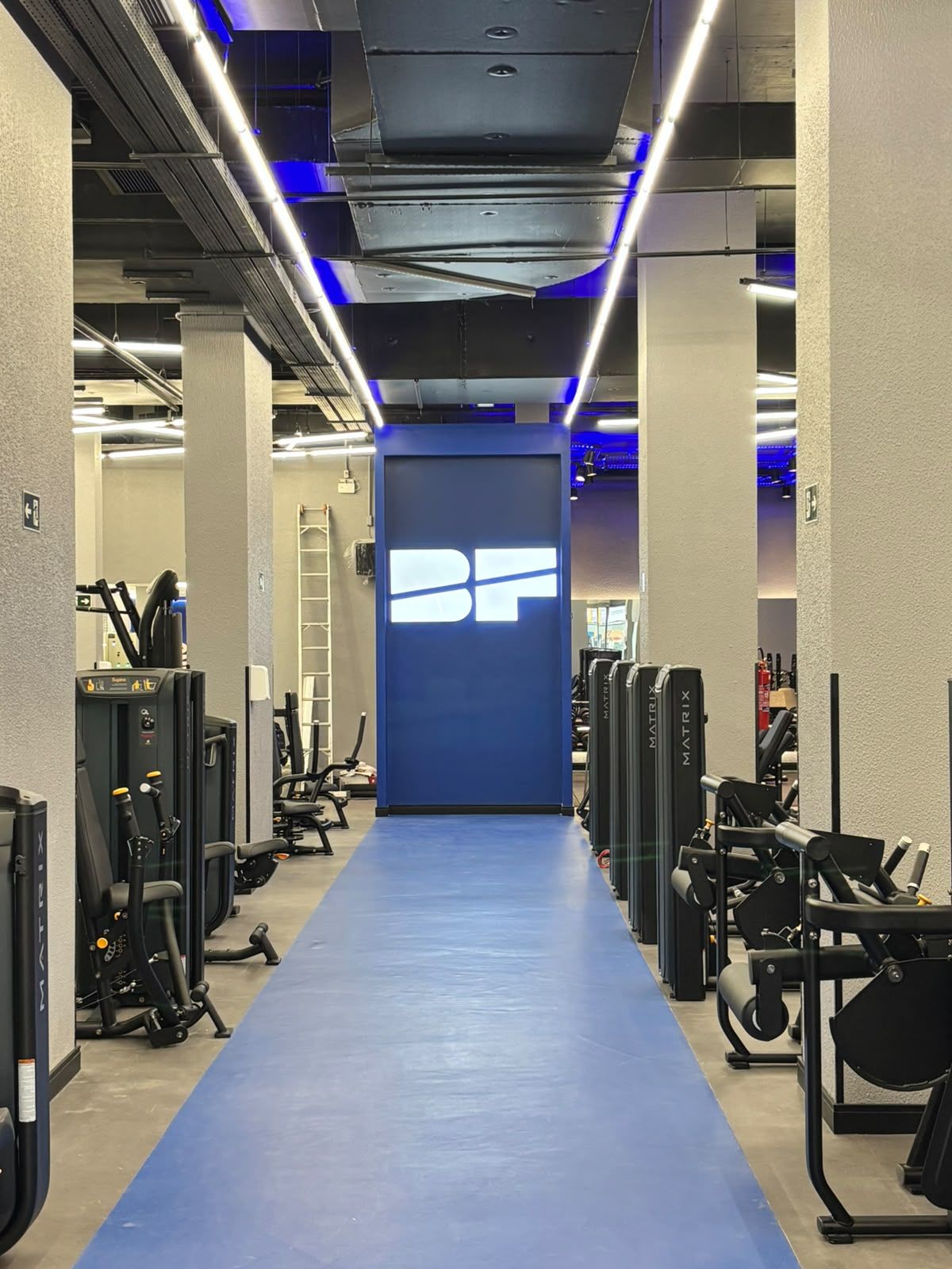 Salão da Bluefit Pedroso de Moraes com corredor central azul, aparelhos de musculação dos dois lados e iluminação linear no teto aparente.