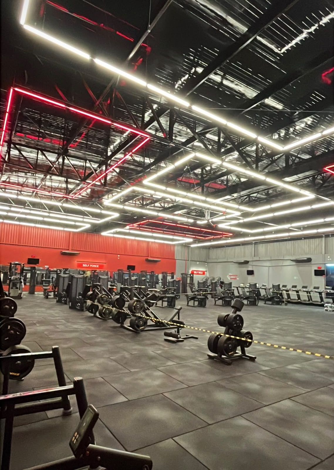Salão da Selfit Aririzal pronto, com piso emborrachado, aparelhos e anilhas, parede vermelha e iluminação em quadros de LED sob a cobertura metálica.